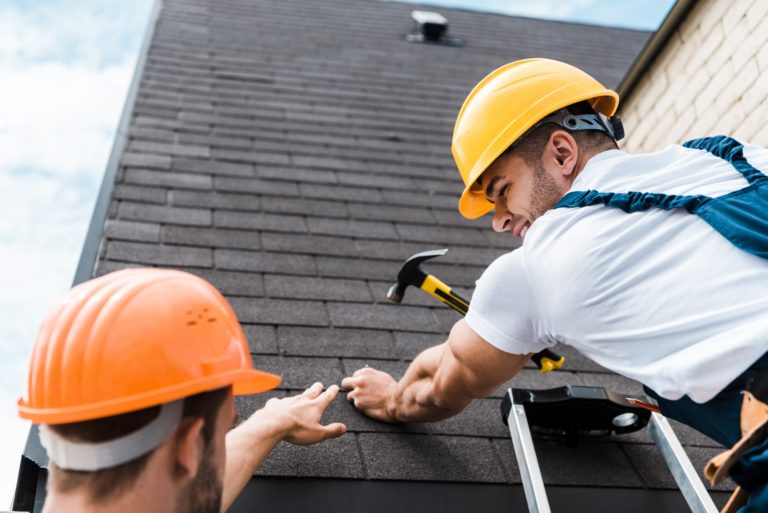 A roofer wearing a hardhat smiling at another worker while hammering a nail into a shingle roof.