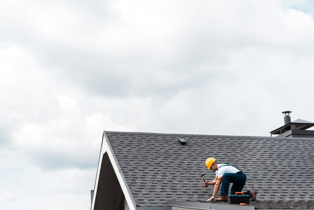 A roofer wearing a hardhat using a hammer on a shingle roof.