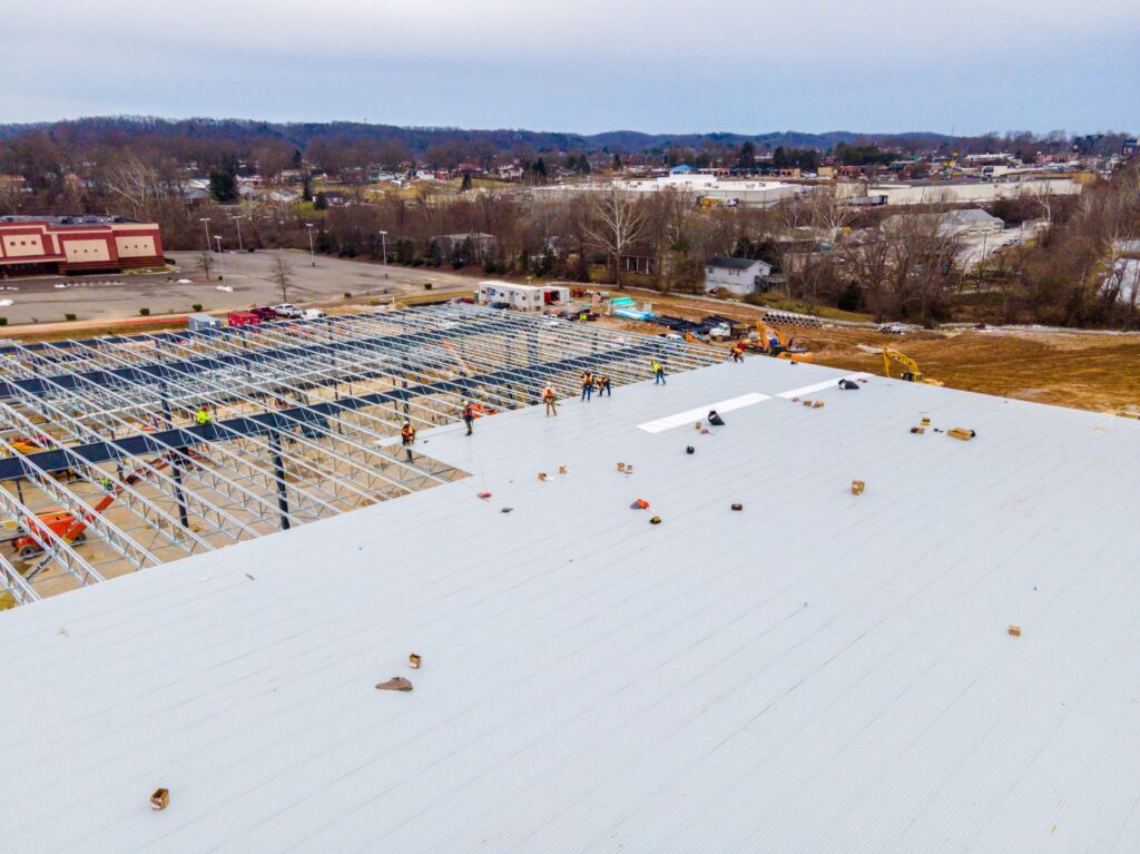 Aerial view of a commercial roofing construction site with workers installing metal panels on a large building framework, surrounded by equipment and materials, with a suburban landscape in the background.