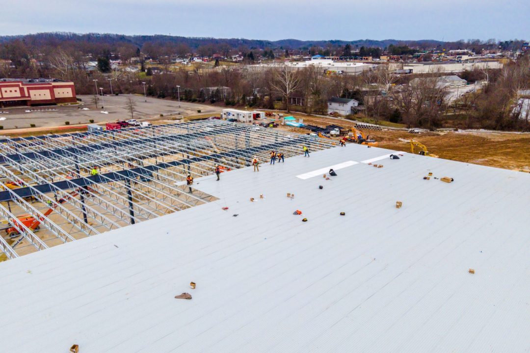 Aerial view of a commercial roofing construction site with workers installing metal panels on a large building framework, surrounded by equipment and materials, with a suburban landscape in the background.
